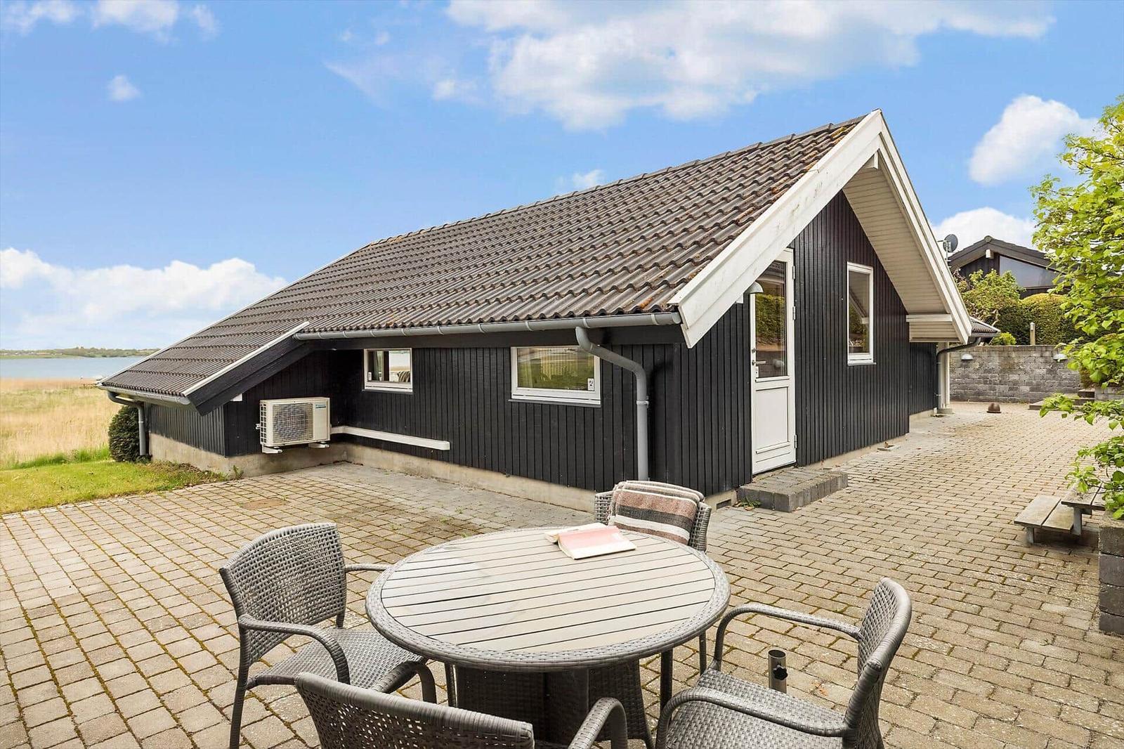 Schwarze Holzhütte mit Terrasse, Tisch und Stühlen. Blick auf Wasser und Himmel.