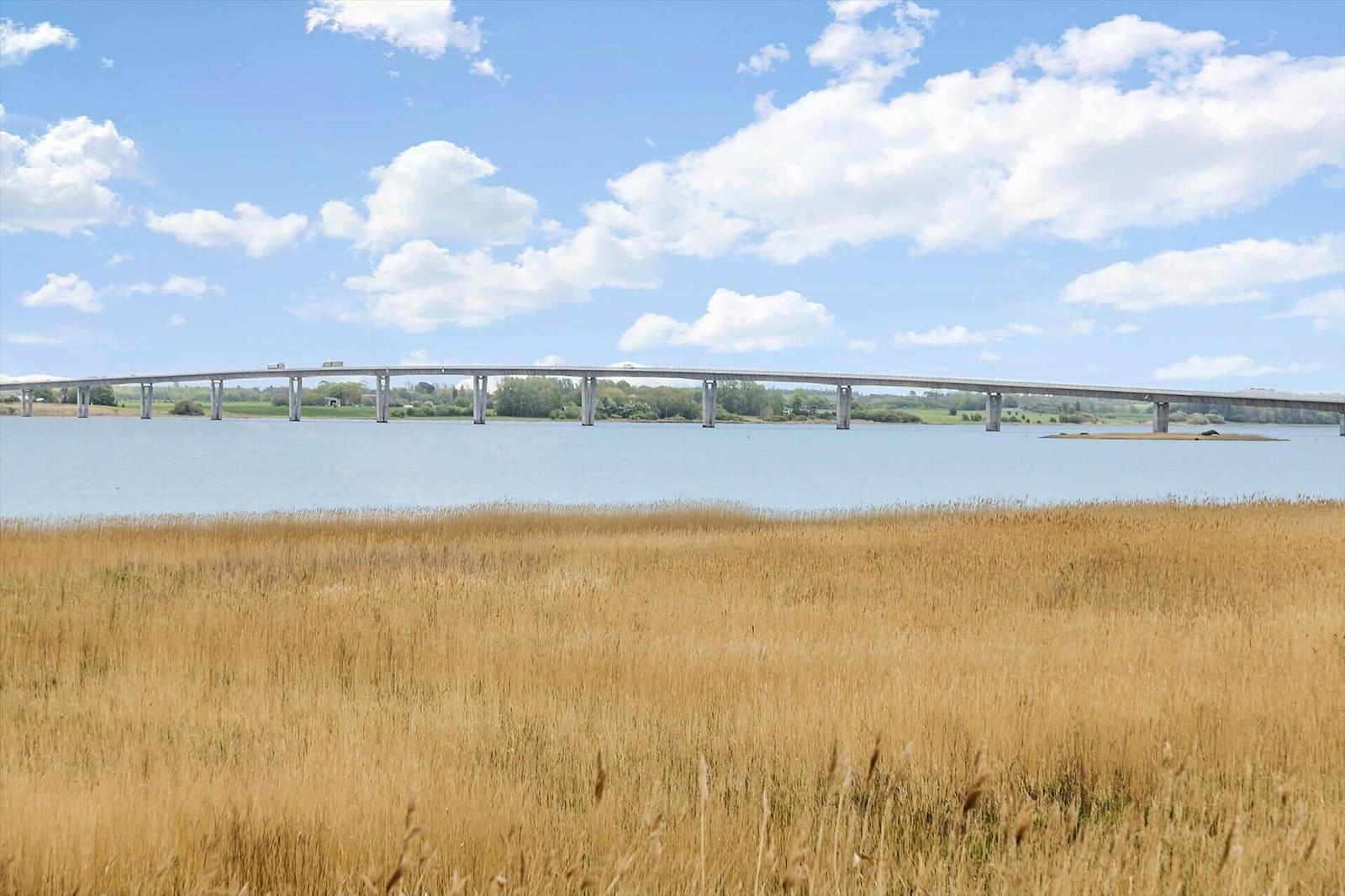 Brücke über einen See mit goldenem Gras im Vordergrund unter blauem Himmel mit weißen Wolken.