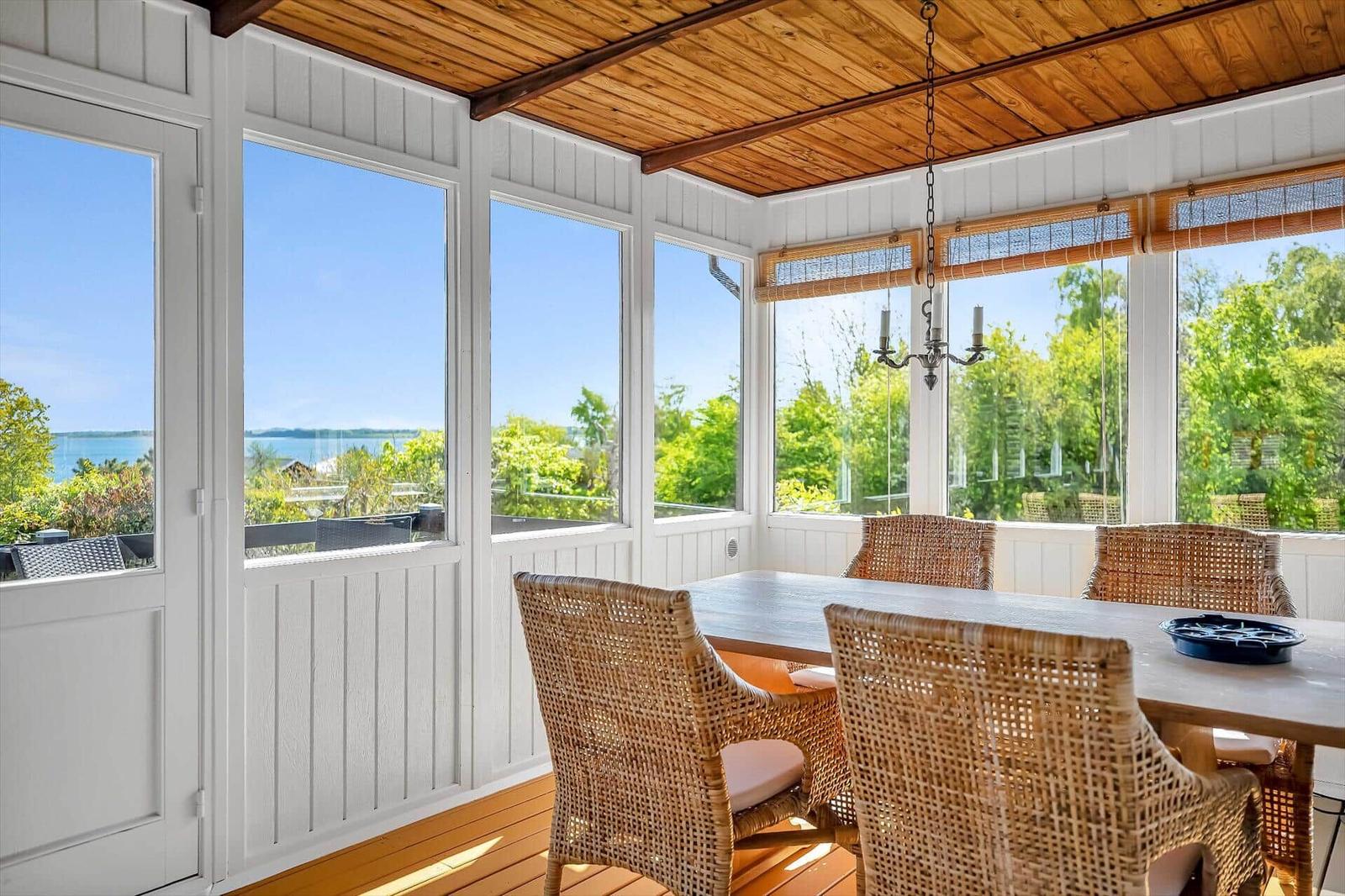 Essbereich mit Sicht auf Wasser und Grün. Holzdecke, wicker Stühle, große Fenster.