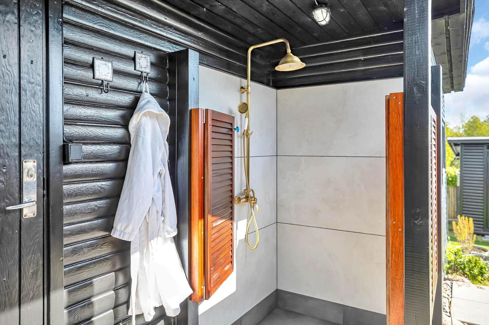 Außen-Dusche mit goldfarbenem Duschsystem und Holzklappe an schwarzer Holzwand.