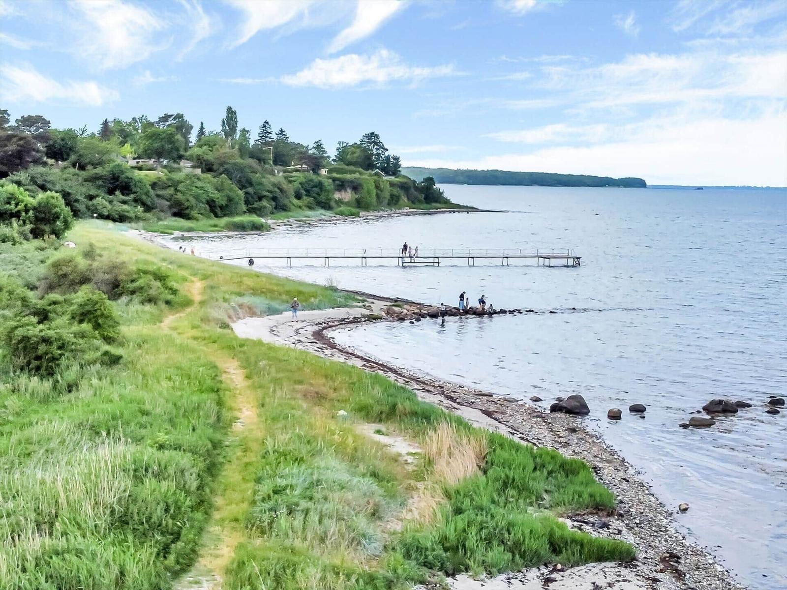 Strand mit Steg und Wanderweg entlang grüner Hanglage.