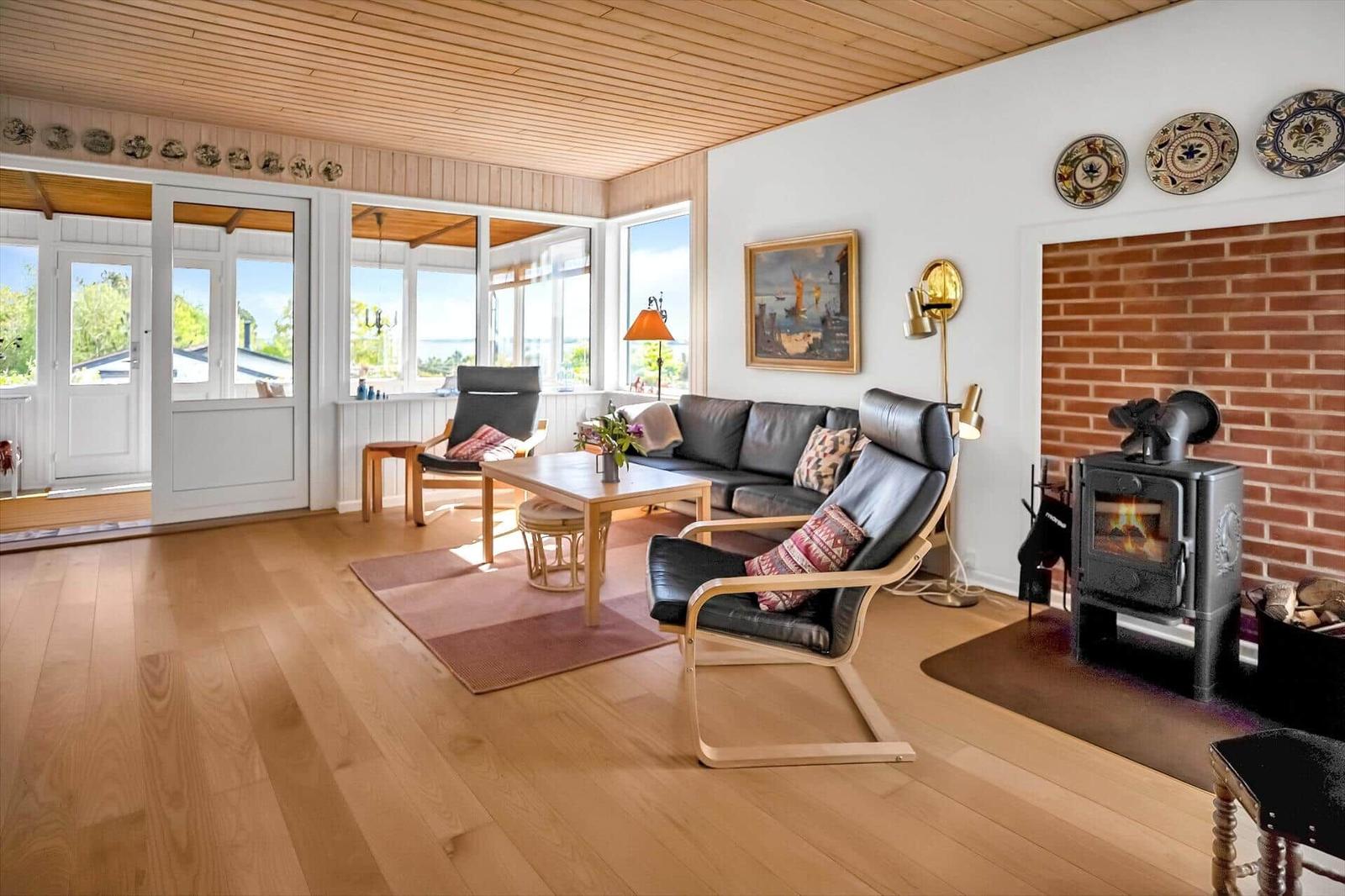 Wohnzimmer mit Holzboden, Sofa, Sessel und Kaminofen. Große Fenster und Tür zum Außenbereich.