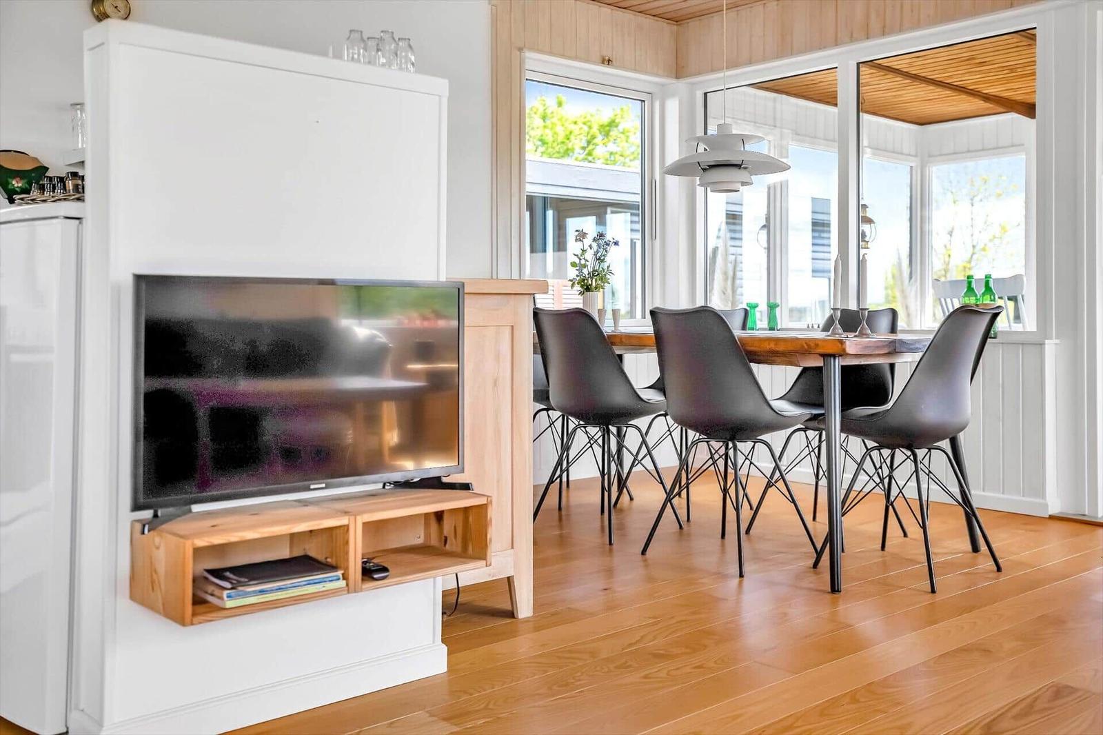 Ein Wohn- und Essbereich mit Fernseher, Holztisch und schwarzen Stühlen. Holzfußboden und große Fenster.