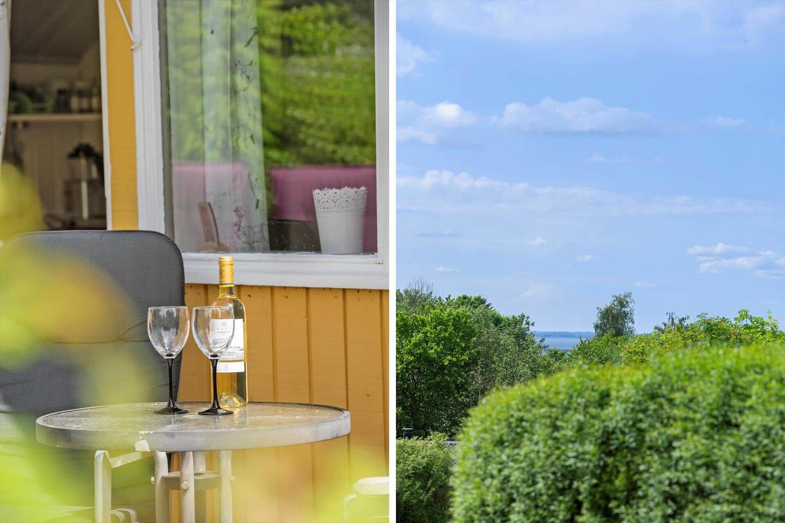Terrasse mit Tisch, zwei Gläsern und einer Weinflasche. Blick auf grüne Bäume und blauen Himmel.