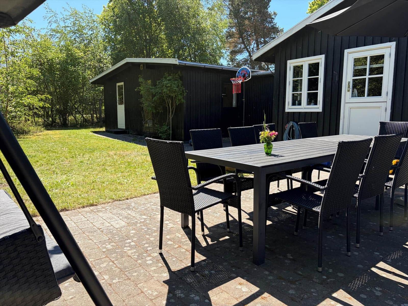 Außenbereich mit Tisch, Stühlen und Grillplatz vor einem schwarzen Holzhaus.