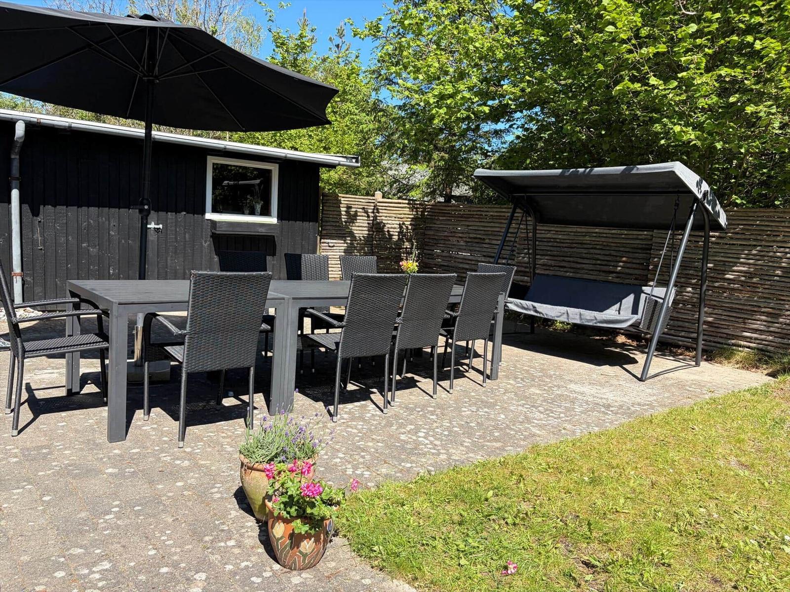 Terrasse mit Tisch, Stühlen, Sonnenschirm und Schaukel unter Bäumen.