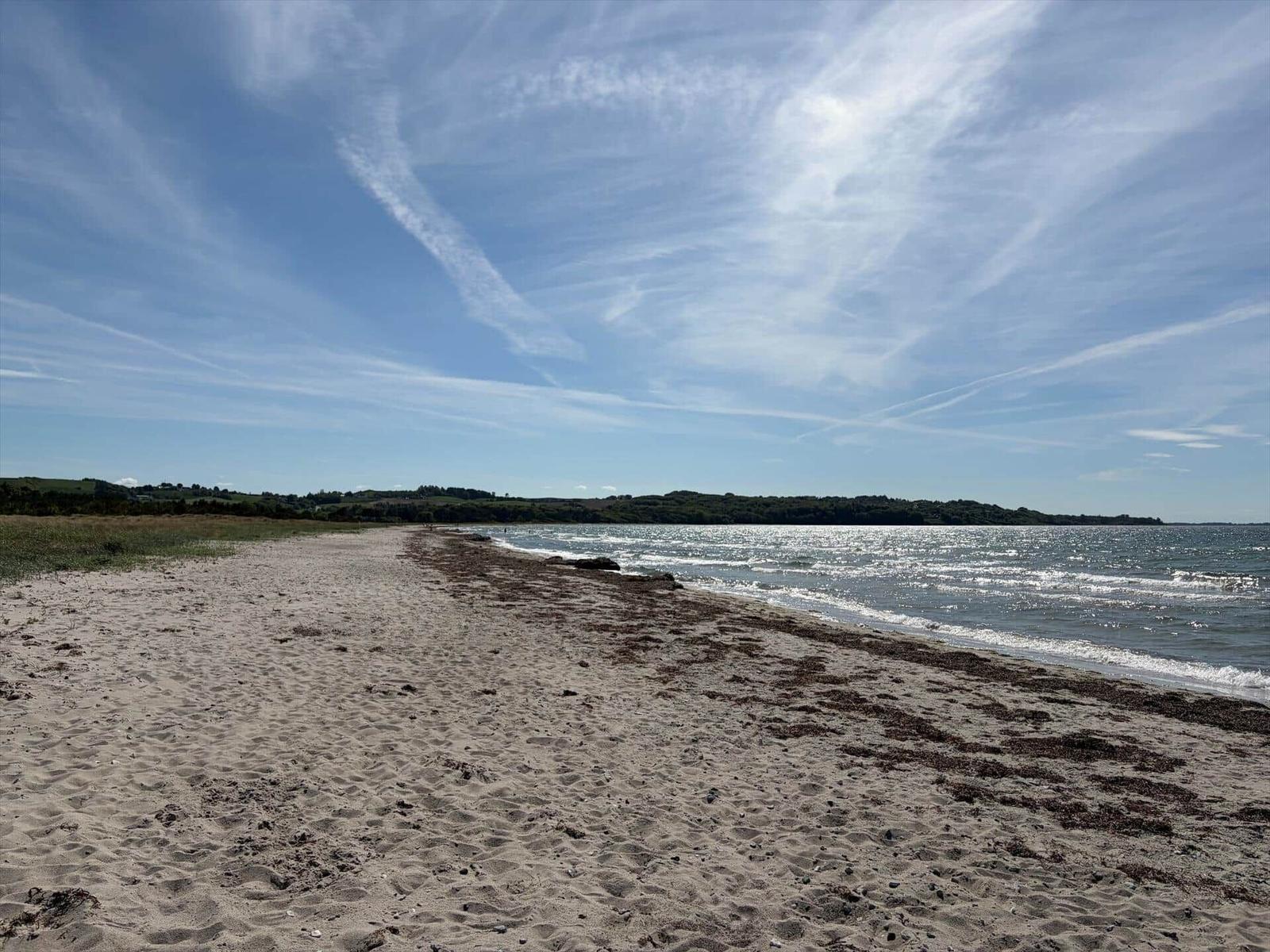 Strand mit Sand und Wellen, Himmel mit weißen Wolken, Ferne mit grünen Hügeln.
