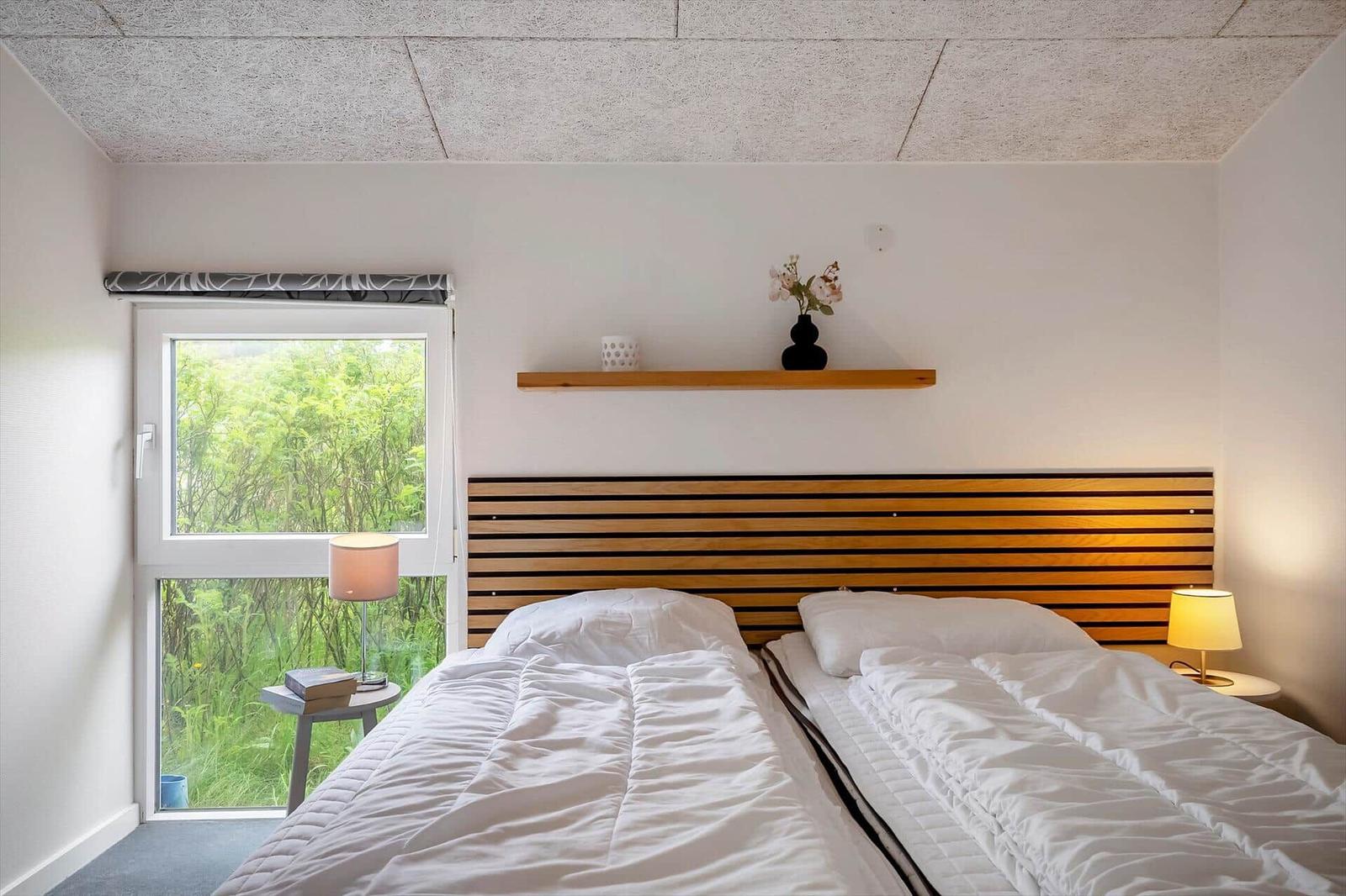 Zimmer mit Doppelbett, Holzkopfteil, Fenster und Nachttisch mit Lampe.