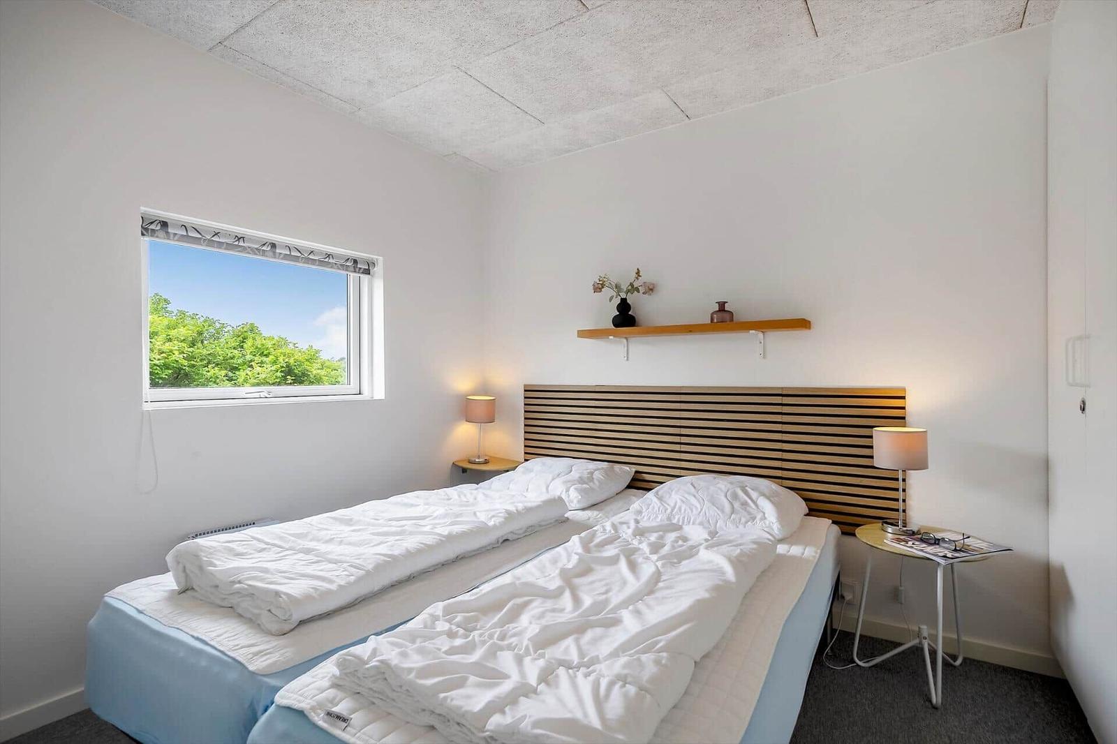 Schlafzimmer mit Doppelbett, Holzkopfteil, zwei Nachttischen und Fenster mit Blick auf Bäume.