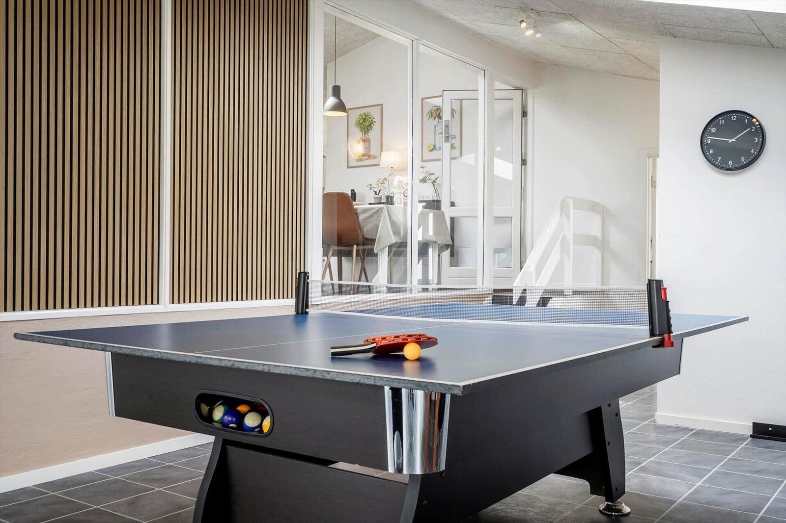 Tischtennisplatte mit Ball und Schläger in einem Raum mit Holzwand und Uhr an der Wand.