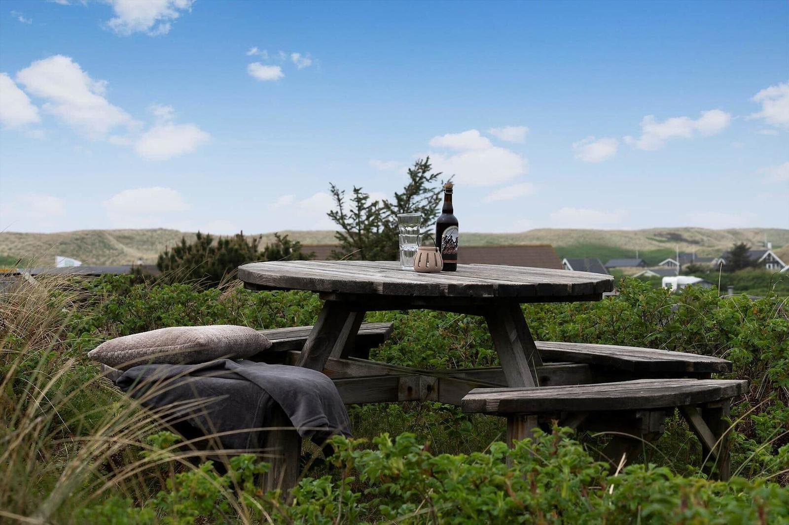 Holztisch mit Flasche und Glas im Grünland. Hintergrund: Hügel und Häuser unter blauem Himmel.