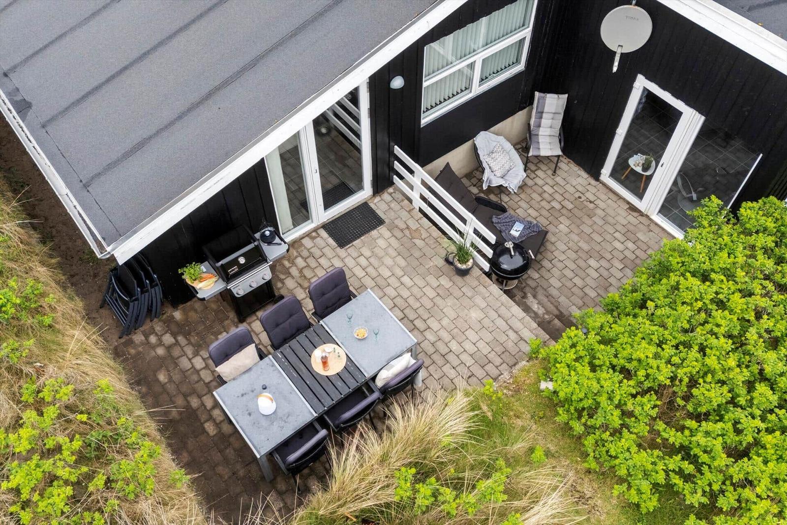 Terrasse mit Grill, Tisch und Stühlen. Schwarze Fassade und Pflanzen umgeben den Außenbereich.