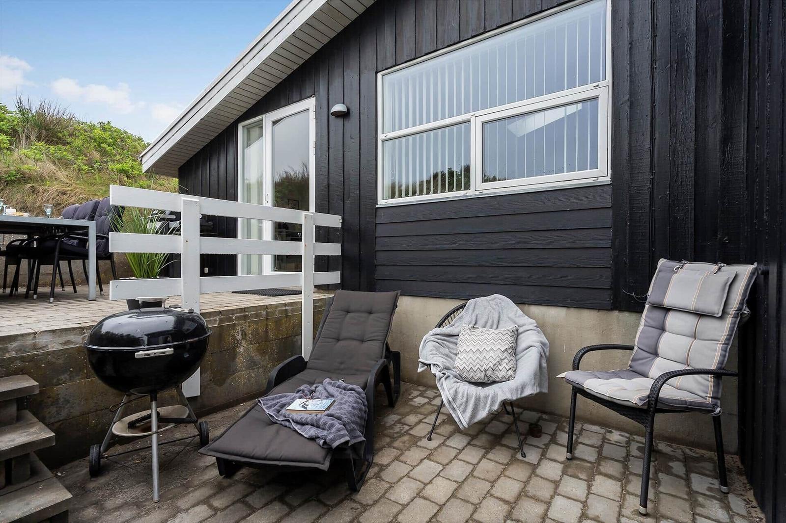 Terrasse mit Liegestuhl, Grill und Sitzgelegenheiten vor schwarzem Holzhaus.