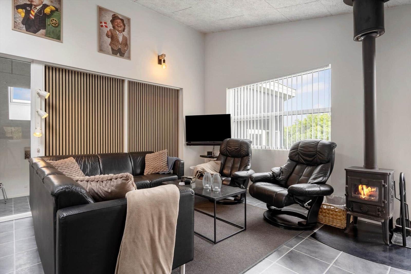 Wohnzimmer mit Sofa, zwei Sesseln, Fernseher und Kaminofen. Große Fenster mit Jalousien.