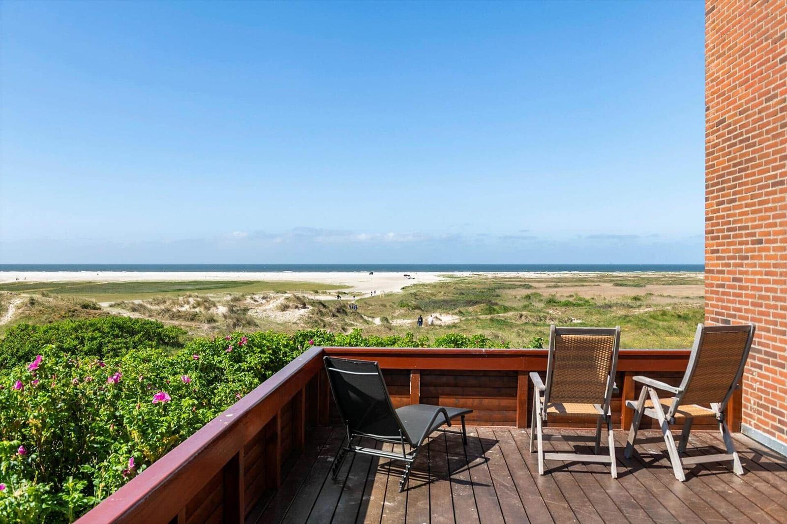 Terrasse mit Liege und Stühlen bietet Blick auf Strand und Dünen.