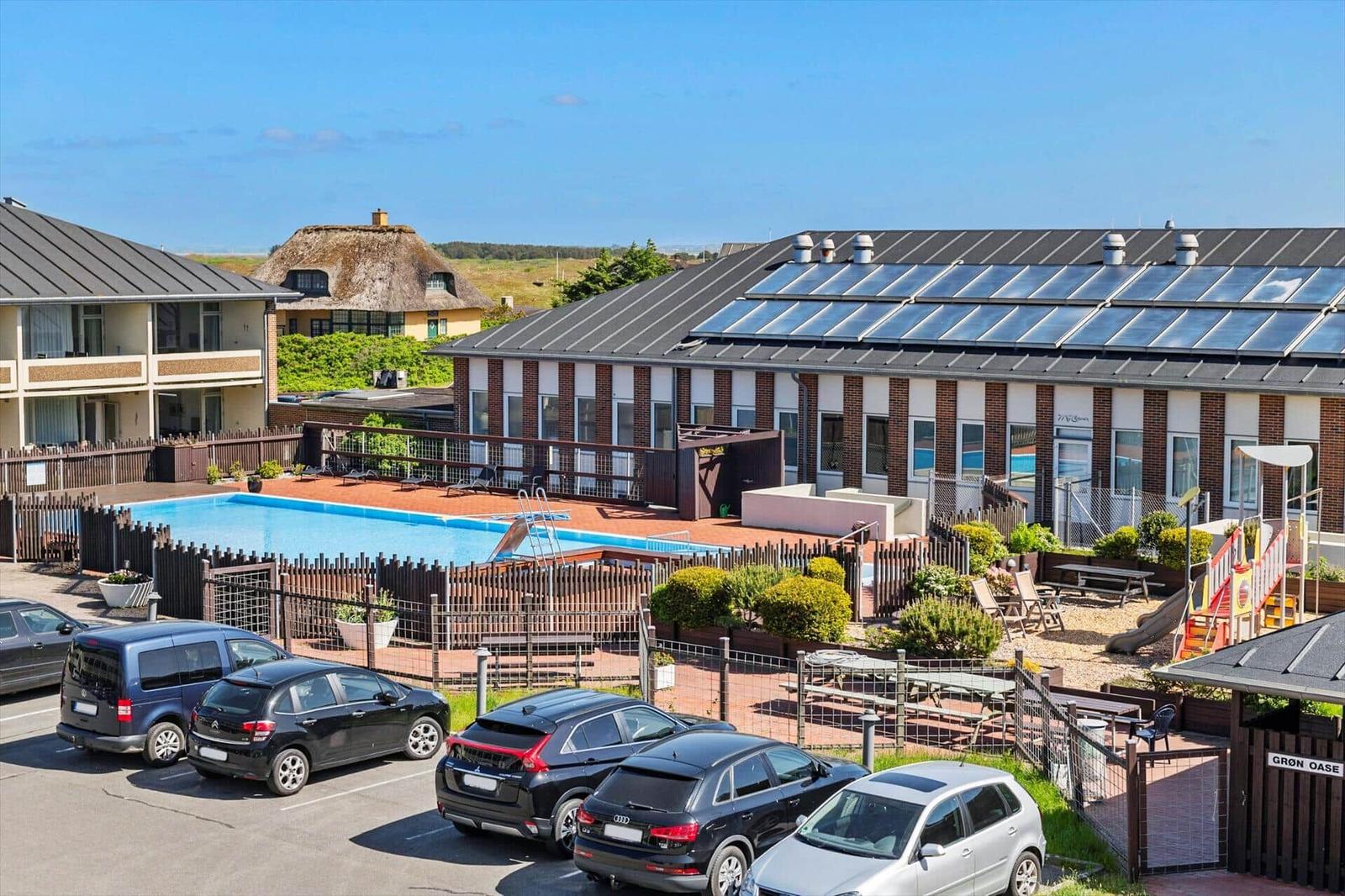Poolbereich mit Sonnenliegen, Spielplatz und Parkplatz vor einem Gebäude mit Solaranlage.