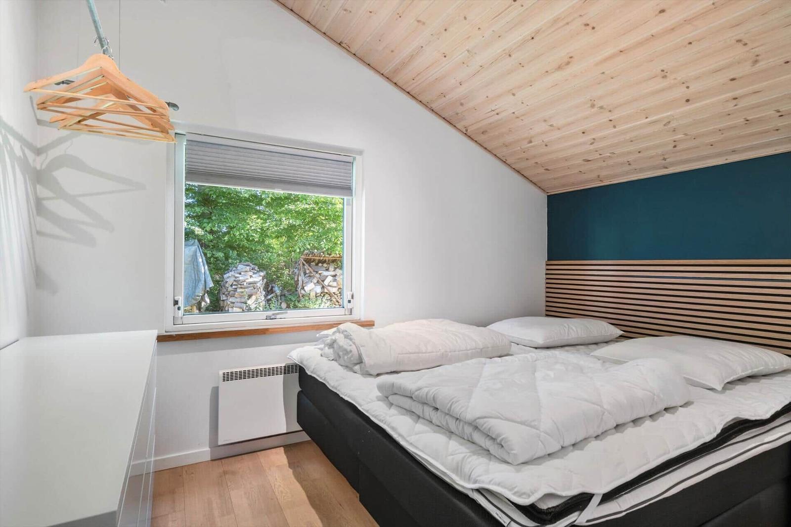 Schlafzimmer mit Bett, Fenster und Holzdecke. An der Wand hängt ein Kleiderhaken.