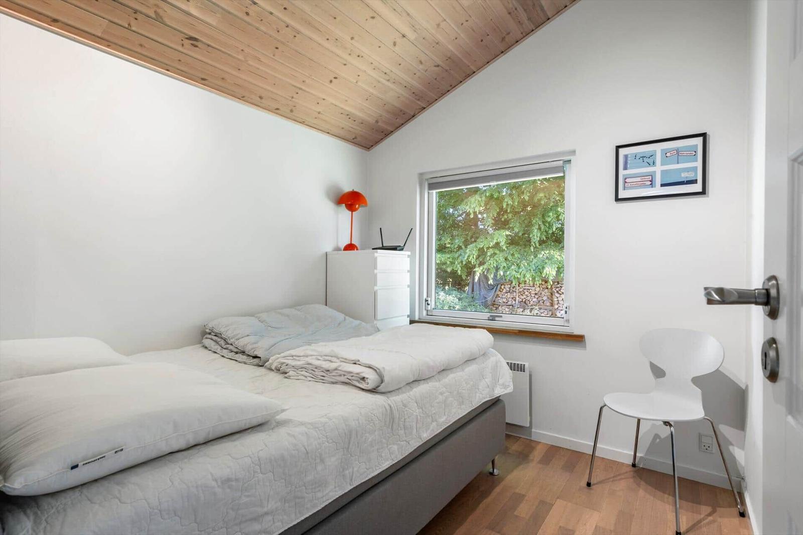 Schlafzimmer mit Bett, Fenster und Stuhl. Holzdecke und weißer Schrank.
