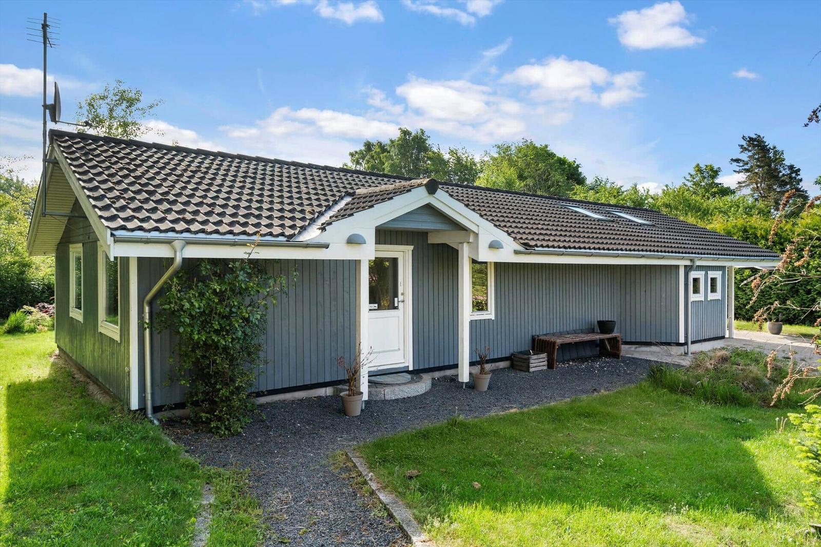 Ein Haus mit grauer Fassade, weißer Tür und Dach mit Schiebedachfenstern. Umgeben von Gras und Bäumen.