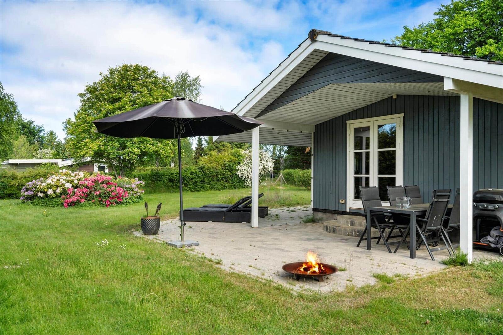 Hütte mit Terrasse, Sitzgruppe, Sonnenschirm und brennendem Feuerkorb im Garten.