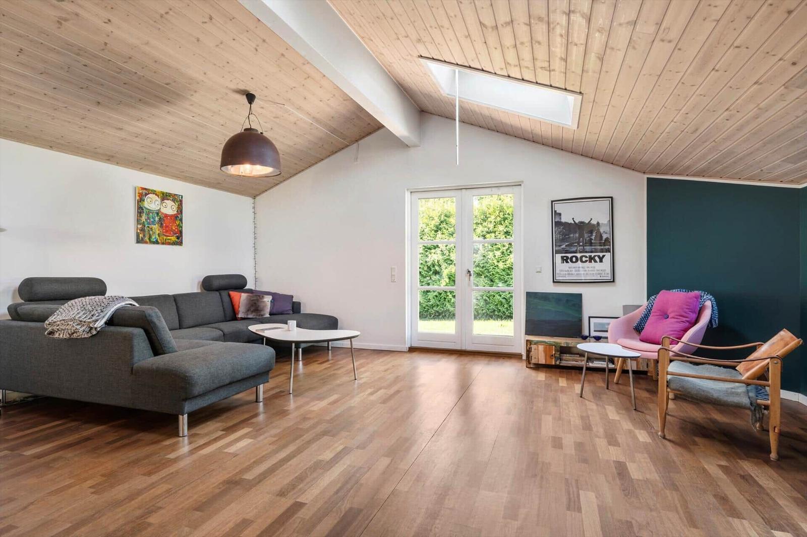 Wohnzimmer mit Holzdecke, Sofa, Sessel und Fenster zur Grünfläche.