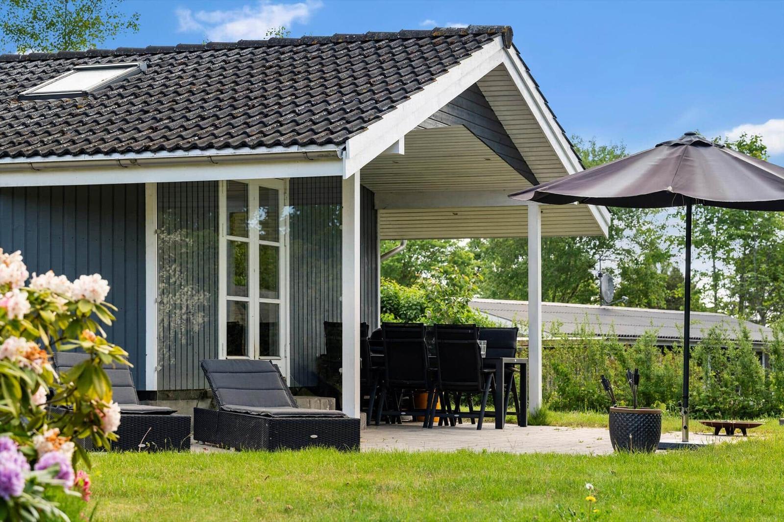Haus mit Terrasse, Sitzgelegenheiten und Sonnenschirm. Grünflächen und Blumen im Vordergrund.