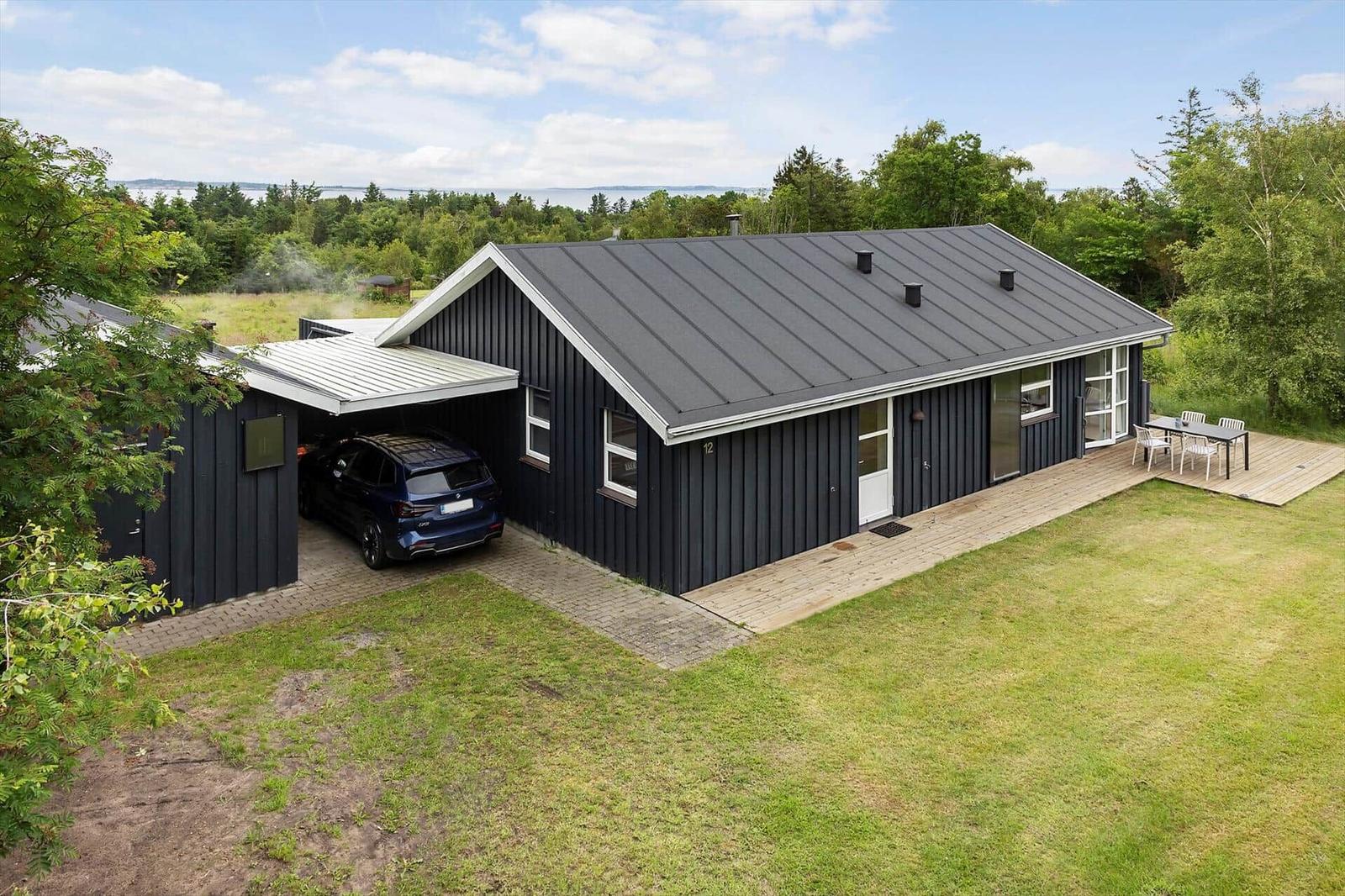 Black house with garage, terrace, and view of forest and water.