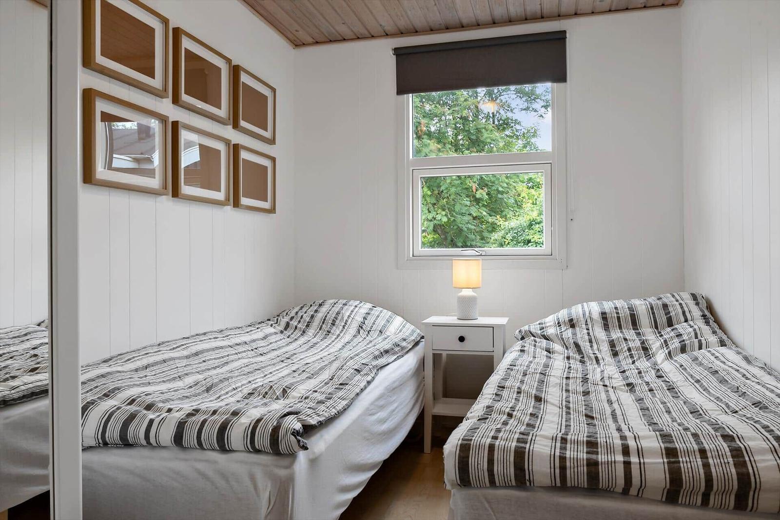 Two beds with striped bedding, a nightstand with lamp, and a window with a view of trees.
