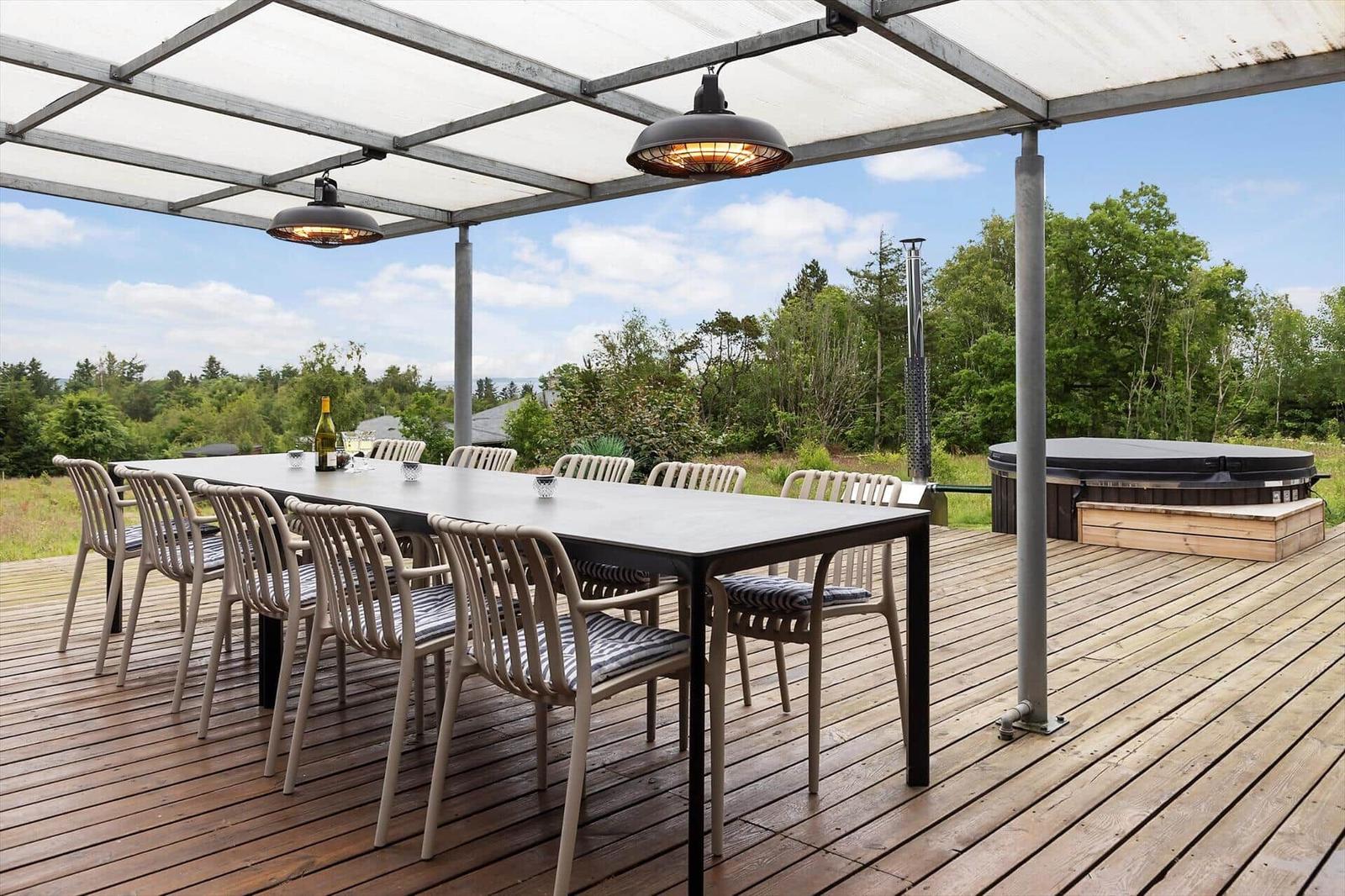 Deck with large table, chairs, and heaters under canopy. Background: forest and hot tub.