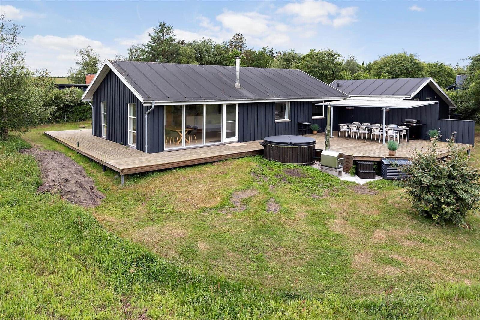 Modern wooden cabin with terrace, hot tub, and grill area in green surroundings.