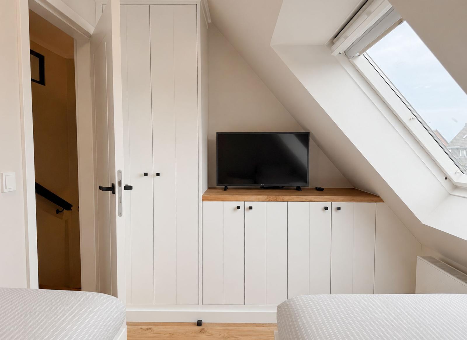 Schlafzimmer mit Dachschrägen, TV und Fenster. Weiße Schränke und Holzboden.