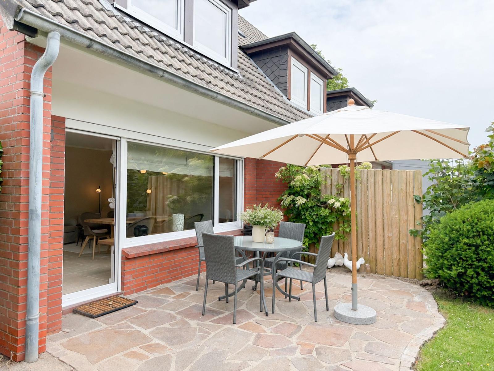 Terrasse mit Tisch, Stühlen und Sonnenschirm. Durchgang ins Haus. Garten mit Hecke und Zaun.