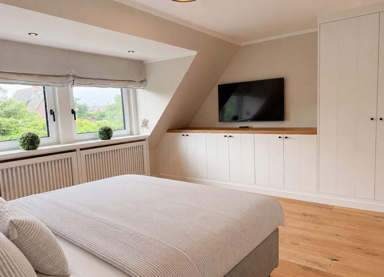Schlafzimmer mit Dachschrägen, TV, Schrank und Fenster mit Blick auf Grün.