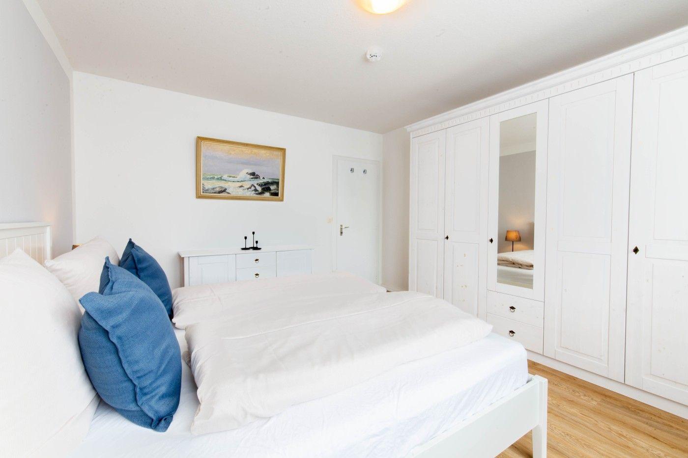 Schlafzimmer mit weißem Bett, blauen Kissen und weißen Schränken. Wandbild und Holzboden.