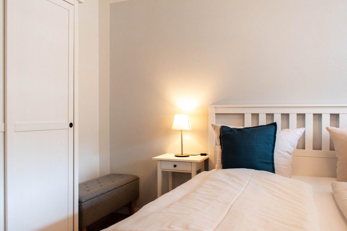 Schlafzimmer mit Bett, Nachttisch und Lampenlicht. Kleiner Sitzhocker und weißer Schrank.