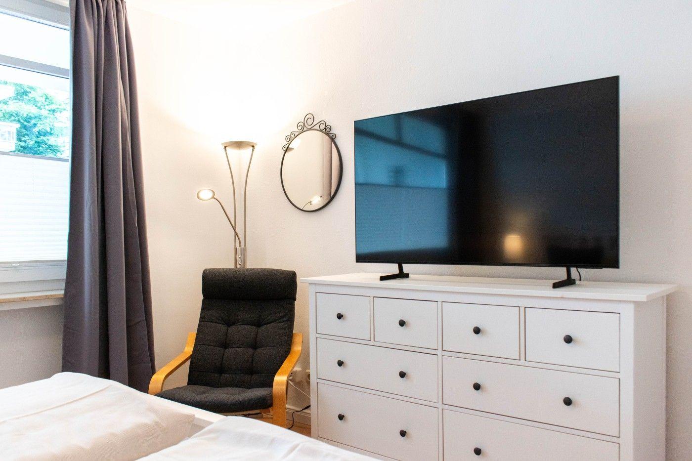 Schlafzimmer mit TV, Schrank und Sessel. Fenster mit Vorhängen.