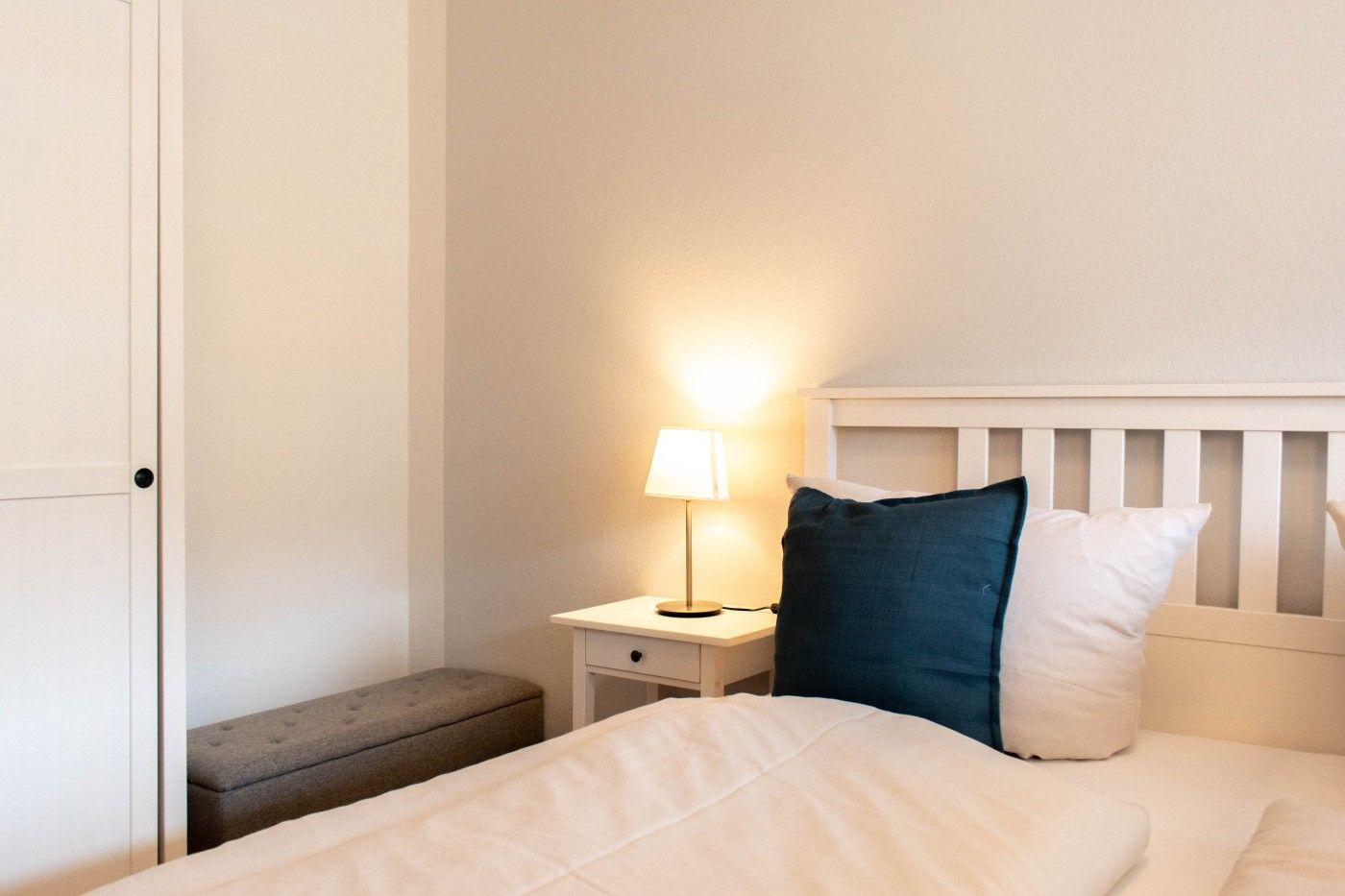 Schlafzimmer mit Bett, Nachttisch und Lampen. Hintergrund ist eine weiße Wand.