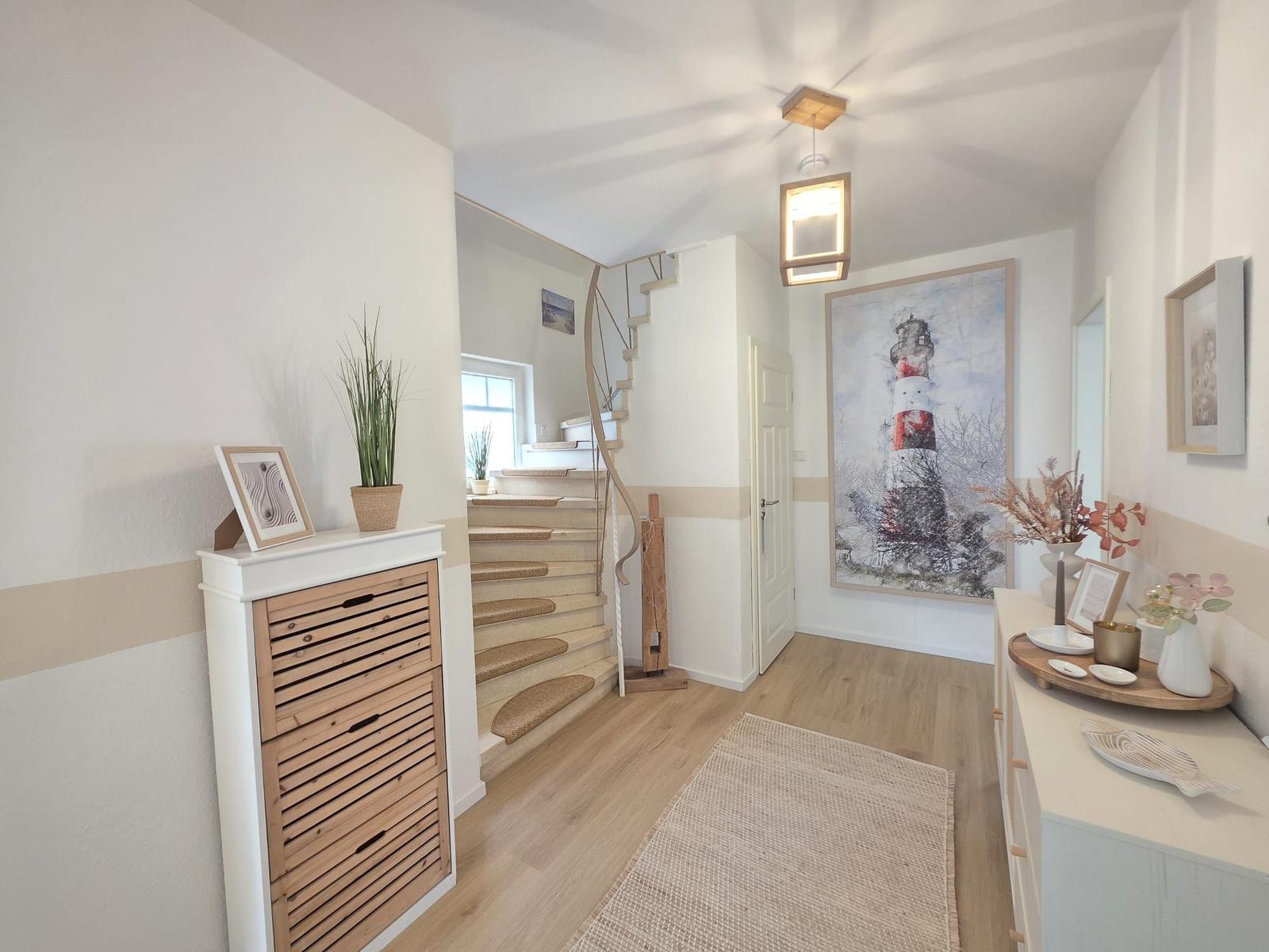 Hausflur mit Holztreppe, Wandbild Leuchtturm und dekorativen Gegenständen auf Möbeln.
