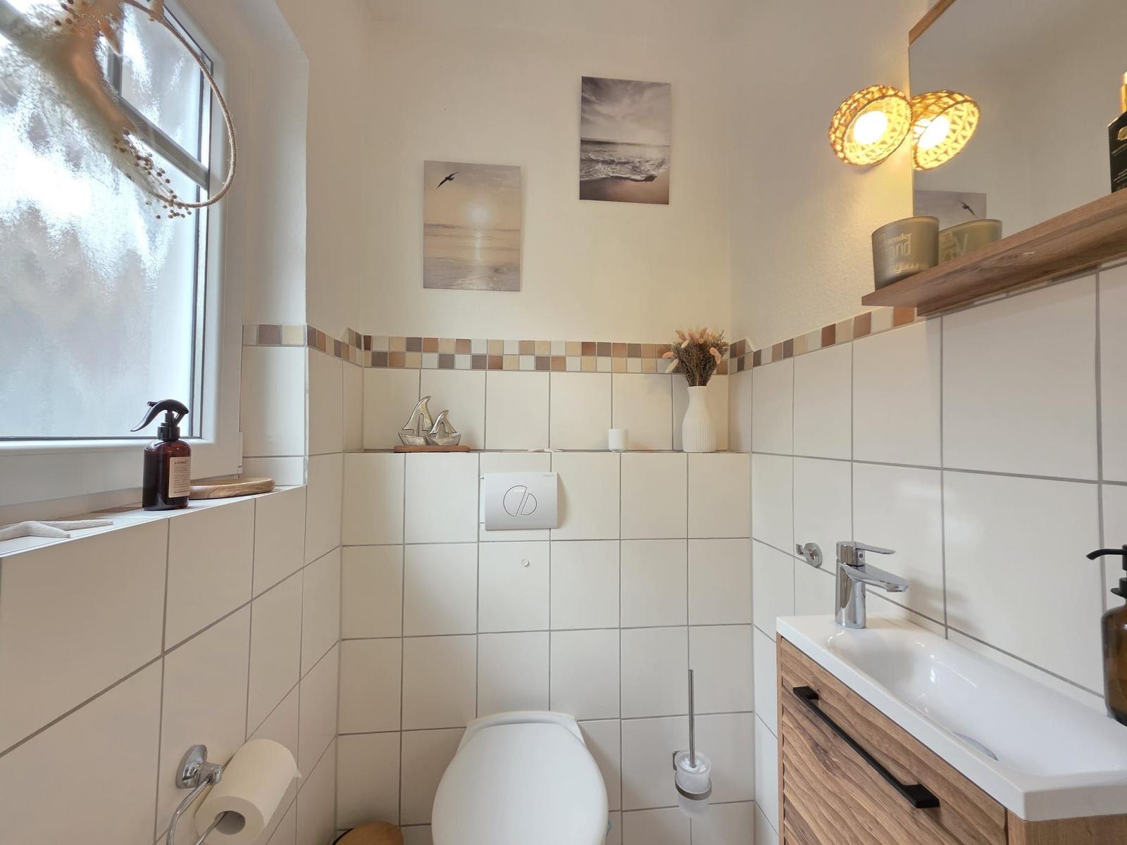 Badezimmer mit Toilette, Waschbecken und Fenster. Wandbilder und Holzregal.