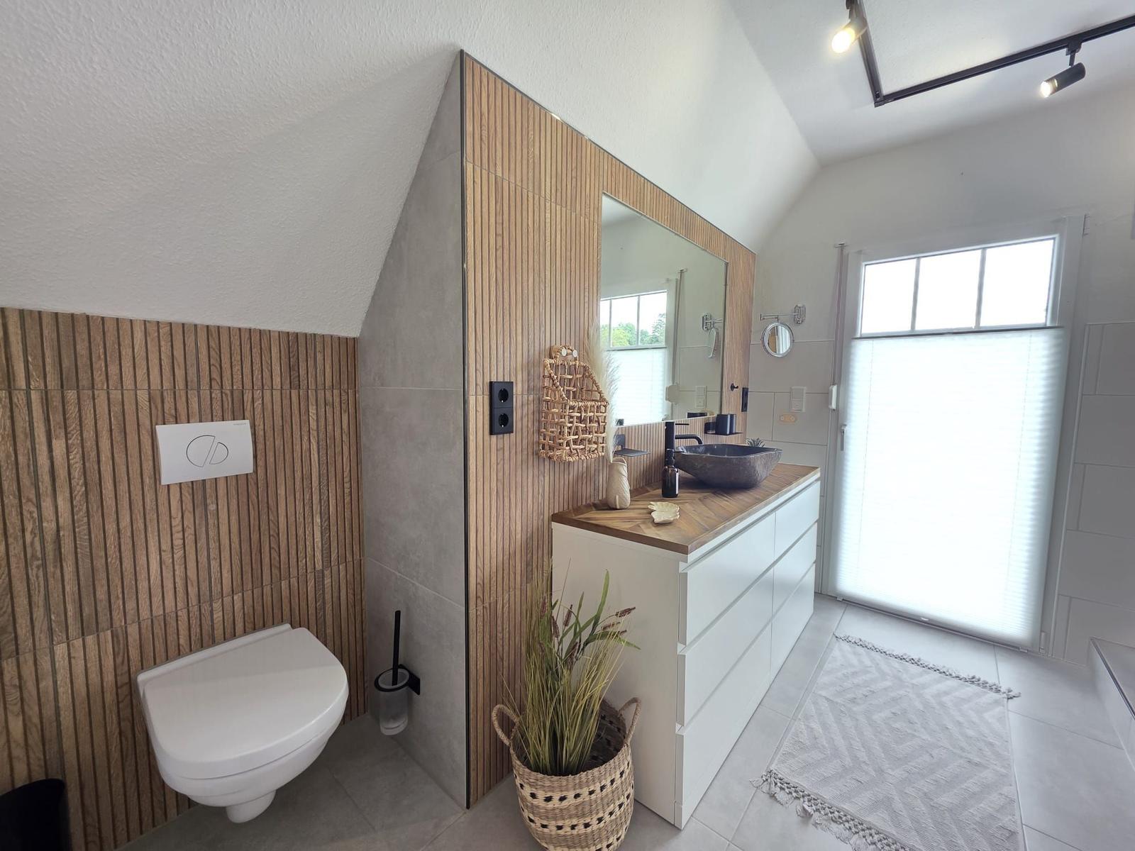 Badezimmer mit Toilette, Waschtisch und Fenster. Holzwand und Spiegel sichtbar.