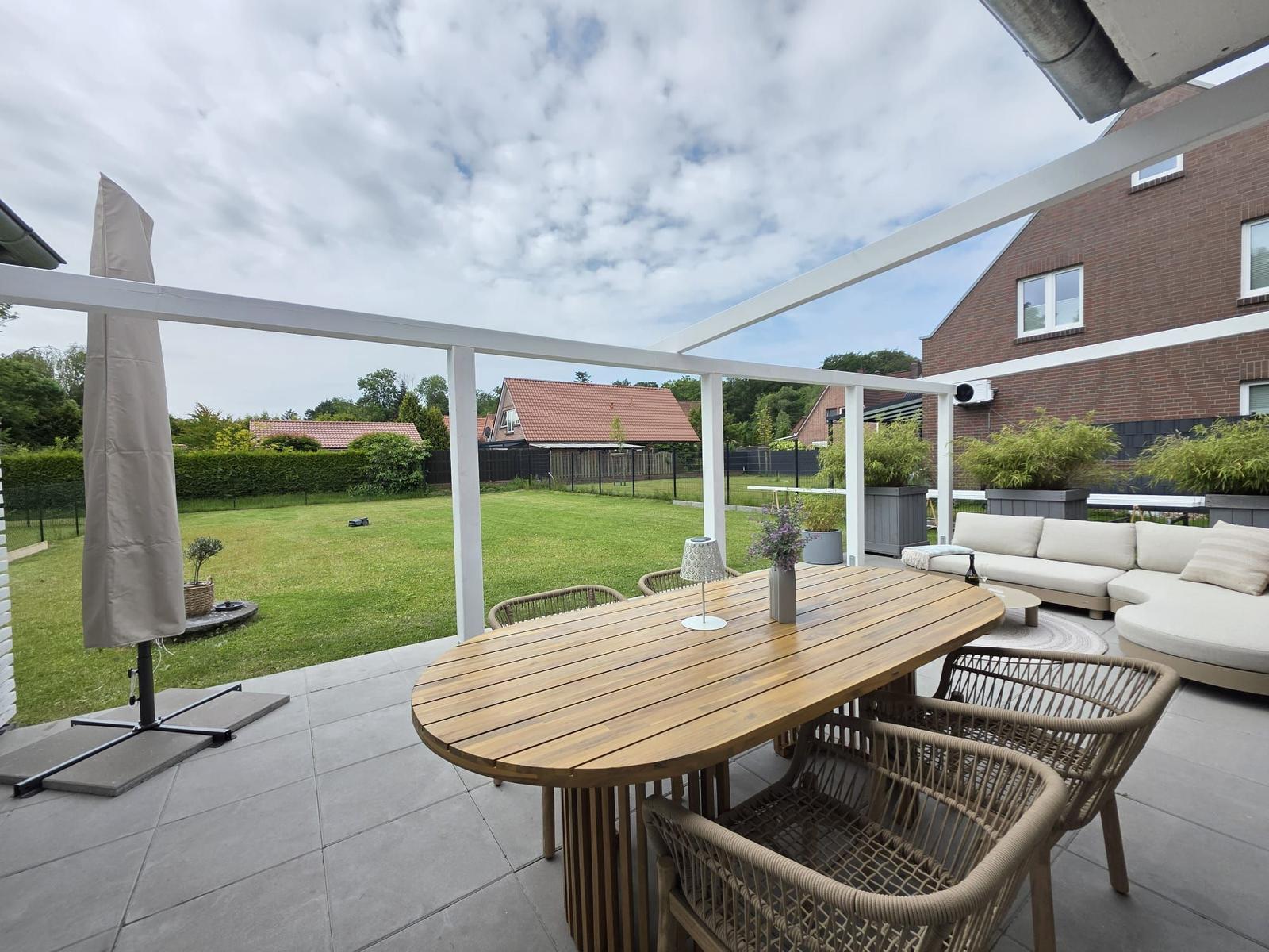Terrasse mit Holztisch, Stühlen und Sofa. Hintergrund: Grasfläche und Nachbarhäuser.