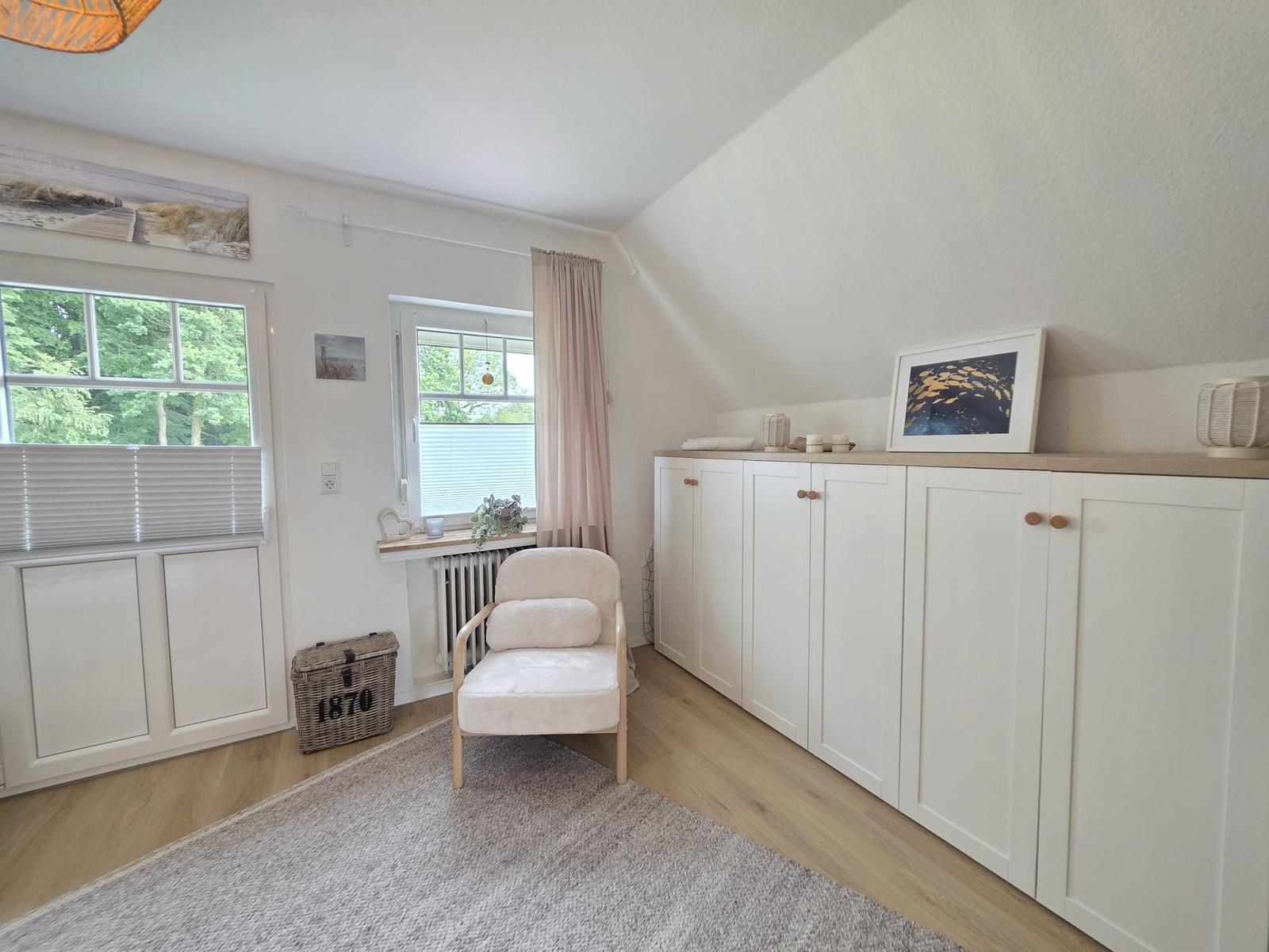 Ein Raum mit weißem Schrank, Sessel und Fenstern mit Blick auf Bäume.