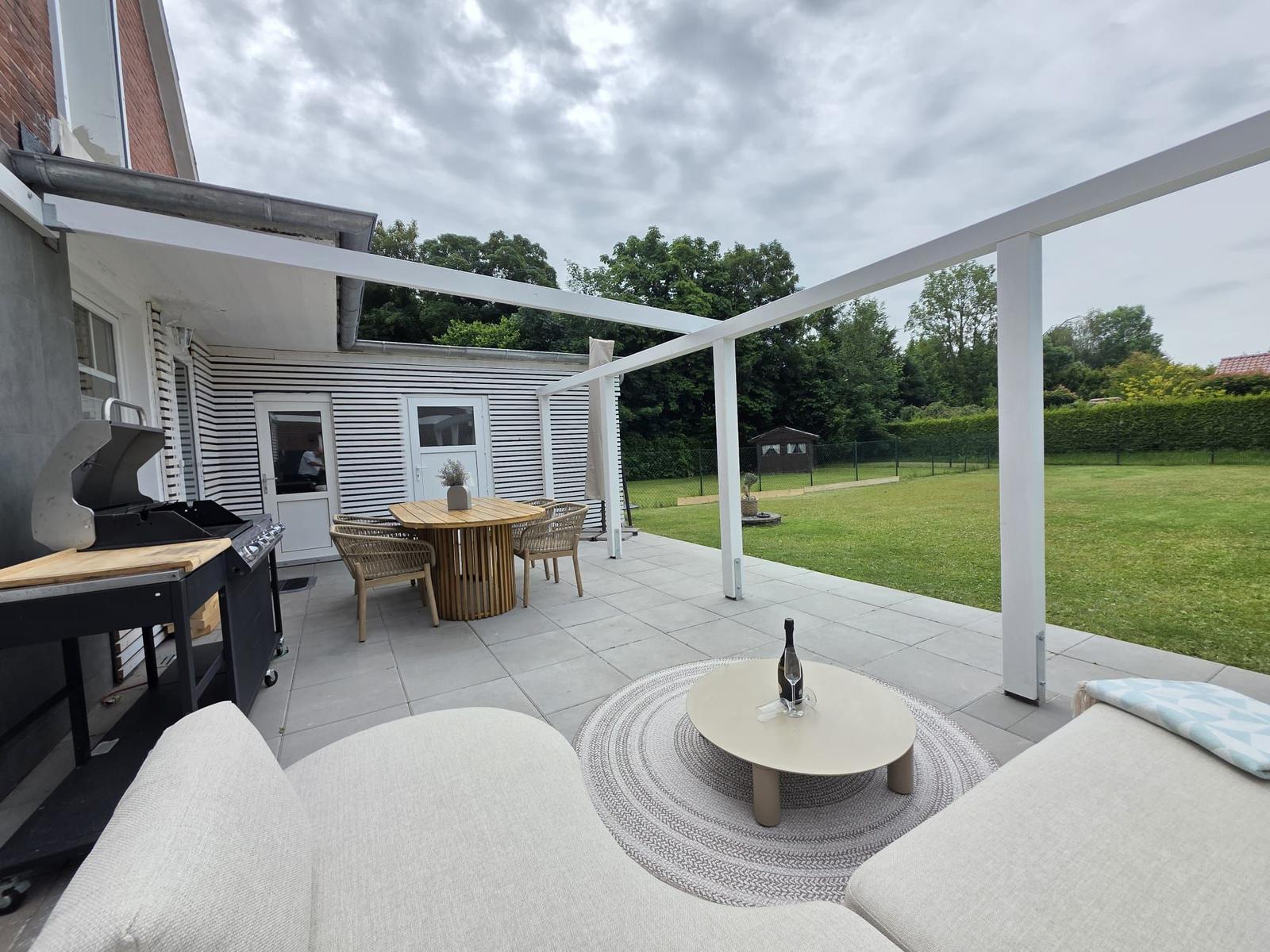 Terrasse mit Sitzgruppe, Esstisch und Grill. Hintergrund: Rasen und Bäume.