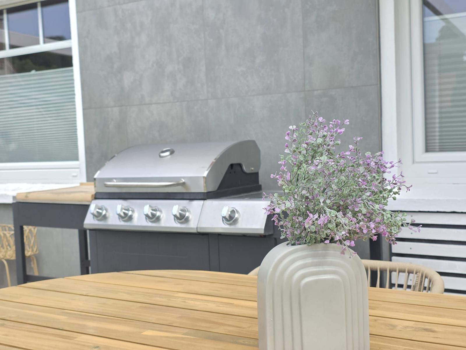 Terrasse mit Grill, Holztisch und Vasenblumen.