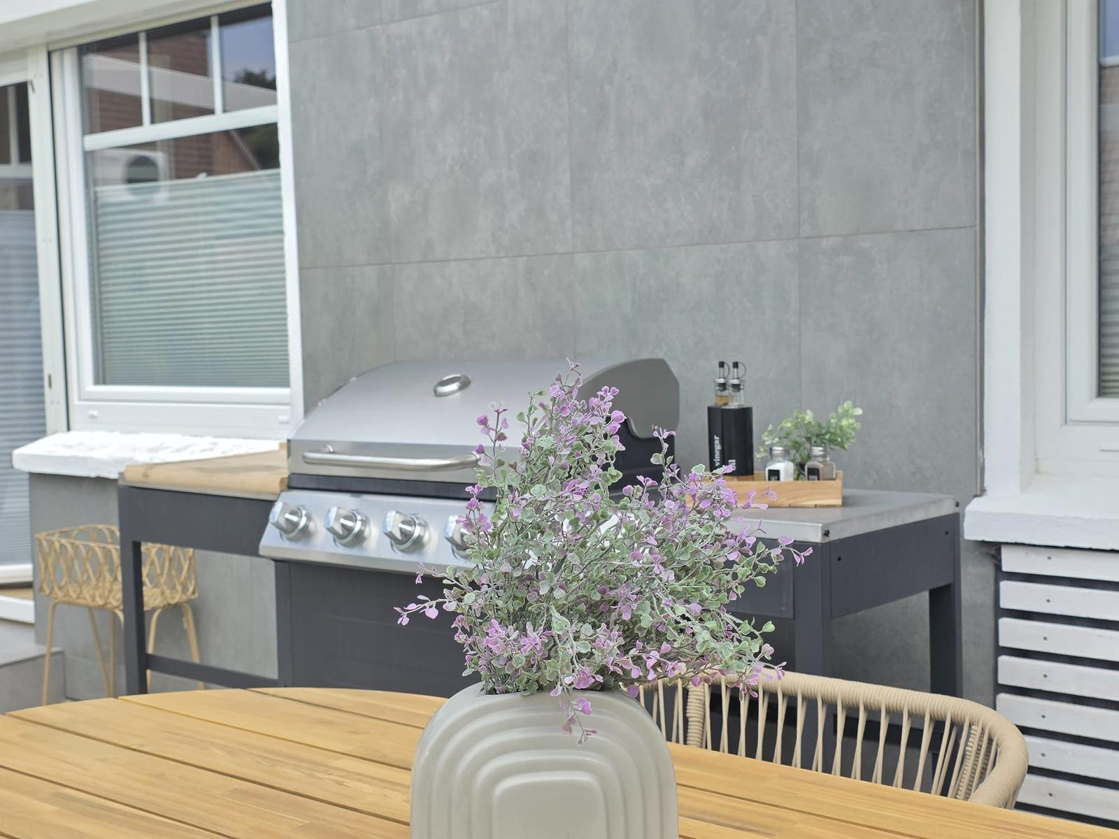 Terrasse mit Grill, Holztisch und Vasenblumen vor grauer Wand.