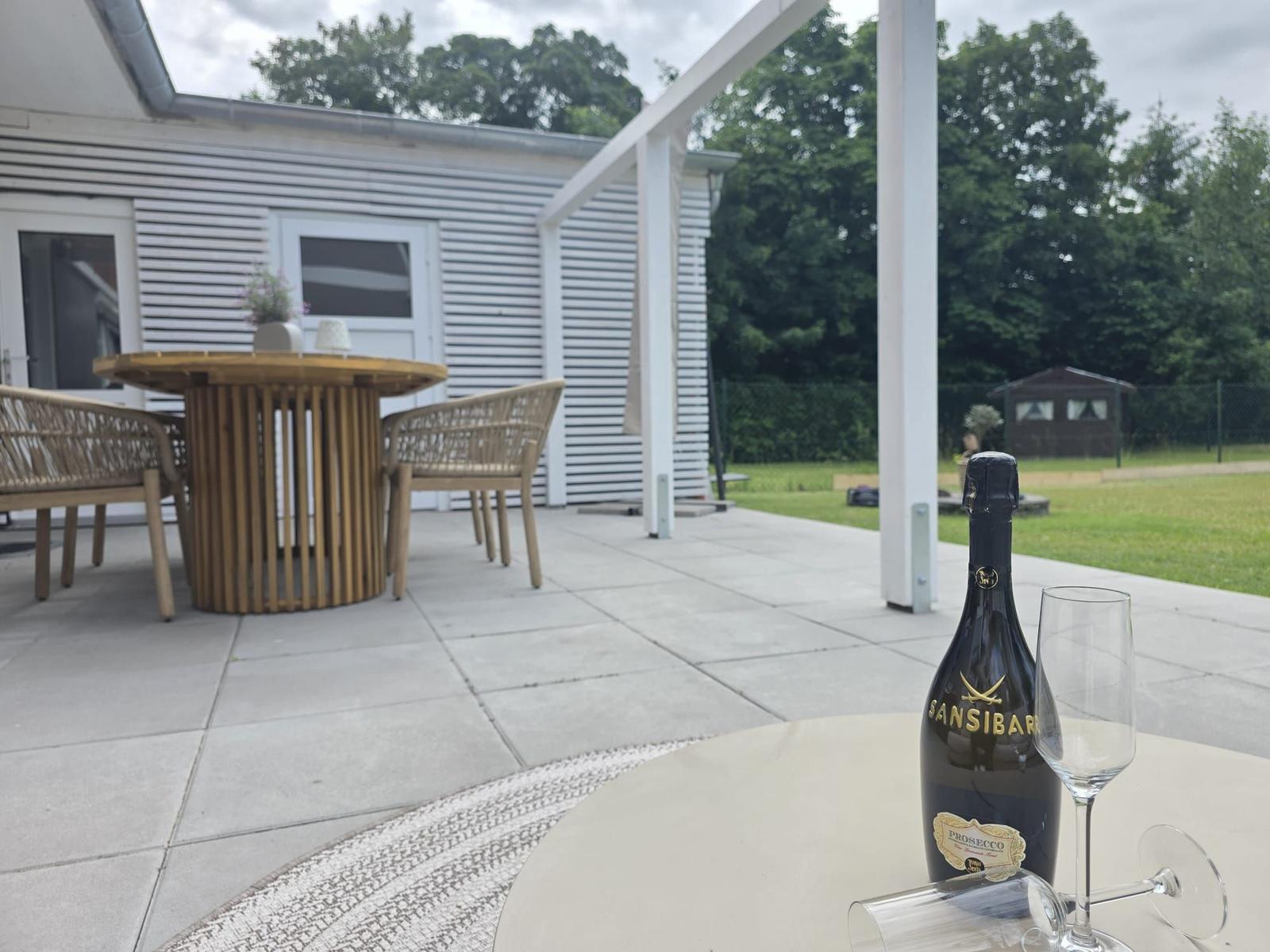 Auf der Terrasse steht ein Tisch mit einer Flasche Prosecco und Gläsern. Im Hintergrund ein weißes Haus mit Garten.