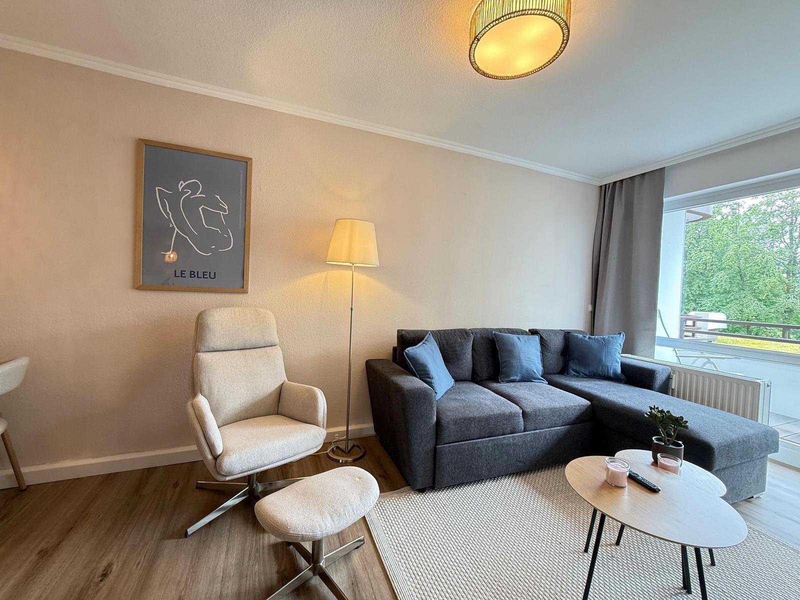 Ein Wohnzimmer mit grauem Sofa, beigefarbenem Sessel und Holzboden. An der Wand hängt ein Bild.