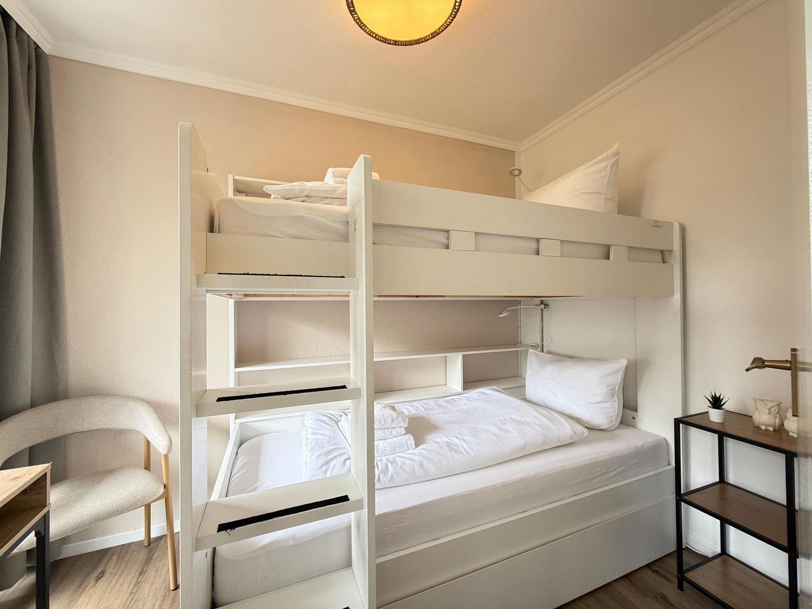Zimmer mit Doppelstockbett, Schreibtisch und Stuhl. Beleuchtung und kleine Regale.