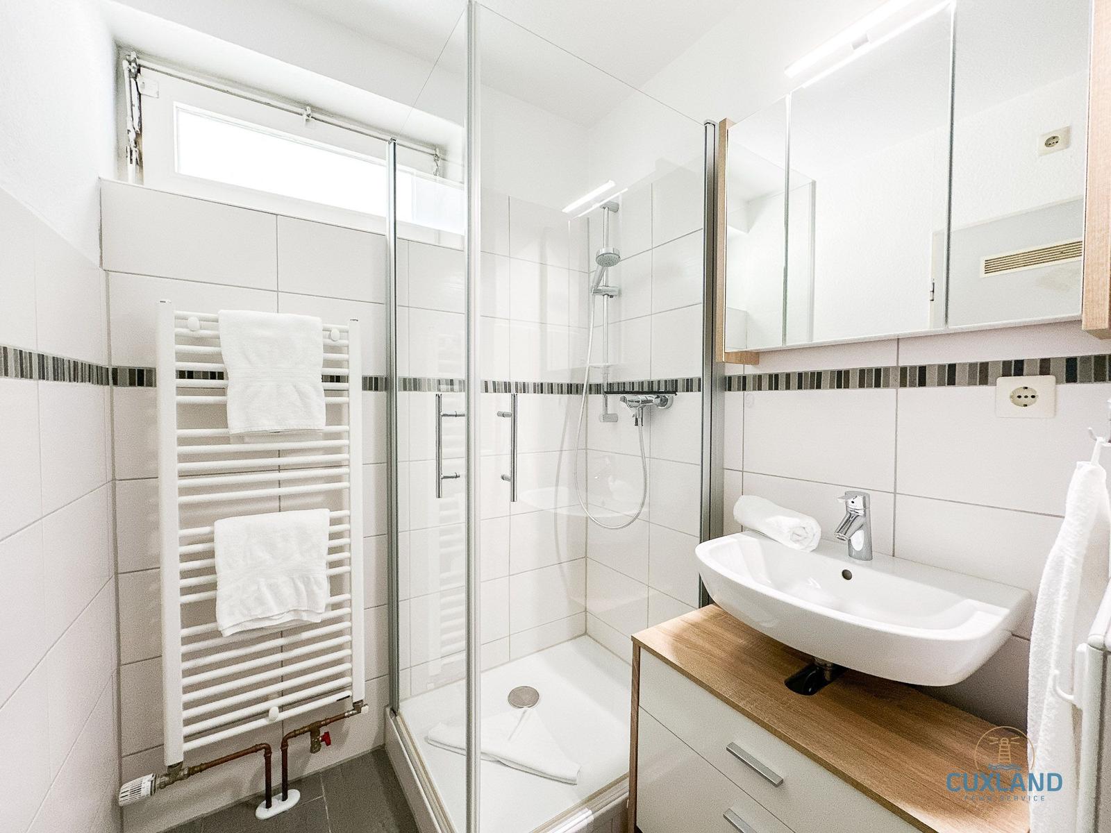 Badezimmer mit Dusche, Waschbecken und Heizung. Weiße Fliesen und Holzoberfläche.