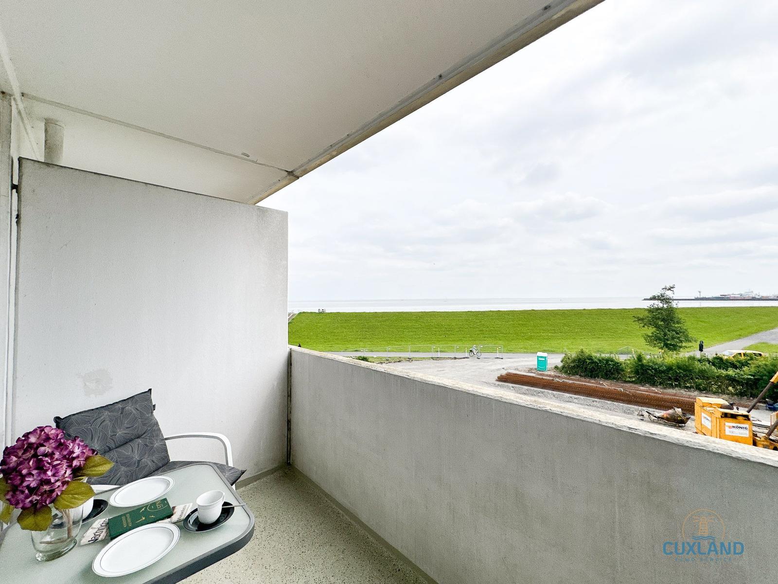 Terrasse mit Tisch, Stuhl und Blumen. Blick auf Grünfläche und Wasser.